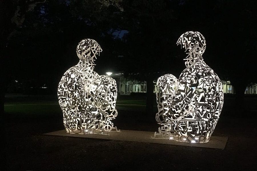 Two Jaume Plensa Mirror sculptures lit from within at night on the Rice University campus — large seated figures composed of openwork steel letters, glowing white against dark trees.