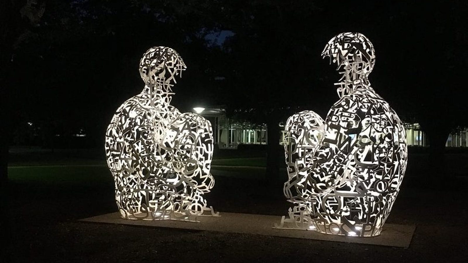 Two Jaume Plensa Mirror sculptures lit from within at night on the Rice University campus — large seated figures composed of openwork steel letters, glowing white against dark trees.