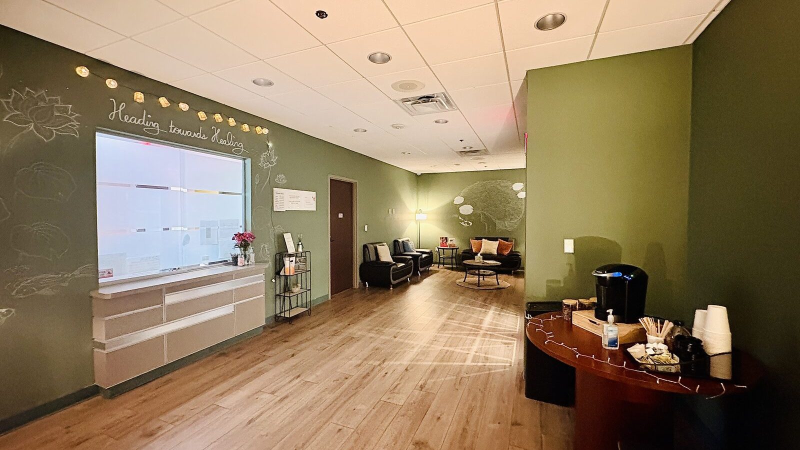 The clinic lounge at Heading Health Stone Oak — green walls, lotus chalk drawings, the message 'Heading towards Healing' written above the reception window, lounge seating, and a coffee setup.